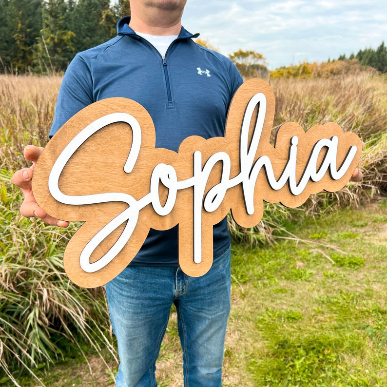 May include: A custom wooden sign featuring the name Sophia in white script lettering mounted on a light brown wooden backing with a smooth finish. The sign has a layered design with the white letters raised above the wooden base.