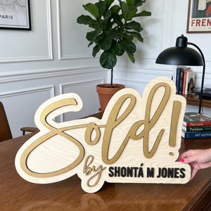 May include: A wooden sign with a light wood grain finish featuring the word Sold! in large, raised gold script lettering. Below, the text by SHONT&Aacute; M JONES is displayed in smaller, raised black lettering on the same light wood base.