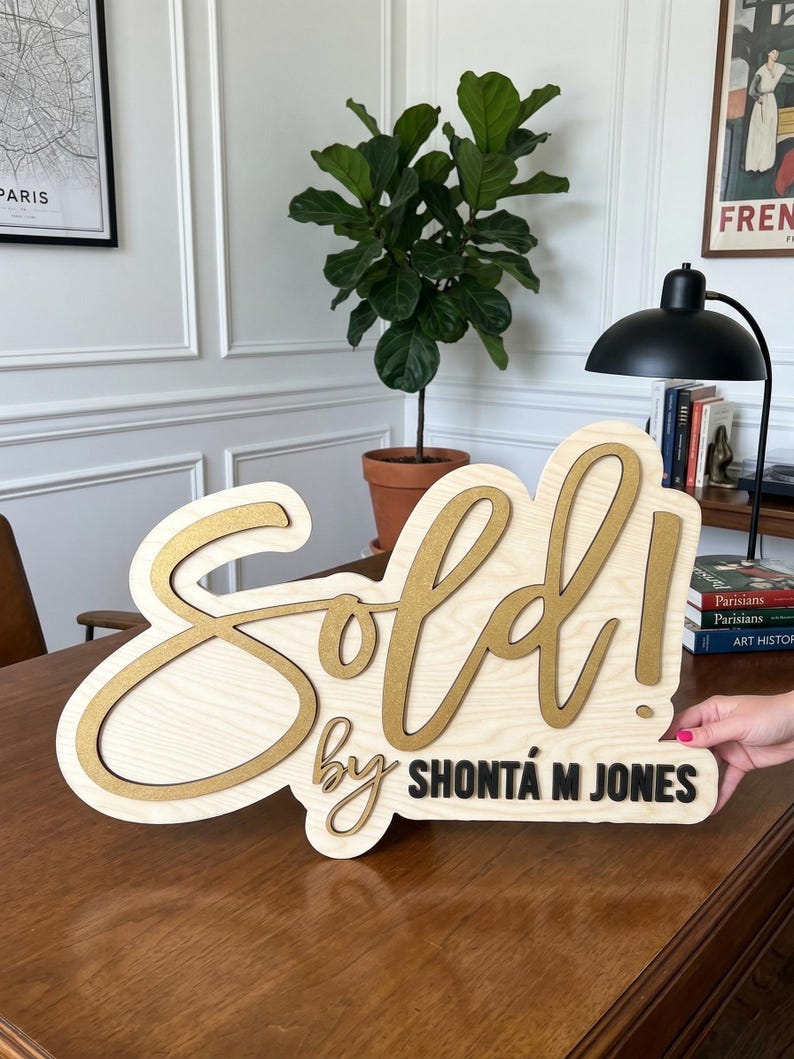 May include: A wooden sign with a light wood grain finish featuring the word Sold! in large, raised gold script lettering. Below, the text by SHONT&Aacute; M JONES is displayed in smaller, raised black lettering on the same light wood base.