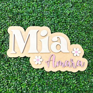 May include: A layered wooden wall sign featuring the name Mia in large white letters and the name Amara in pink cursive script. The design includes two small white flowers with pink centers, all mounted on a light-colored wooden backing board.