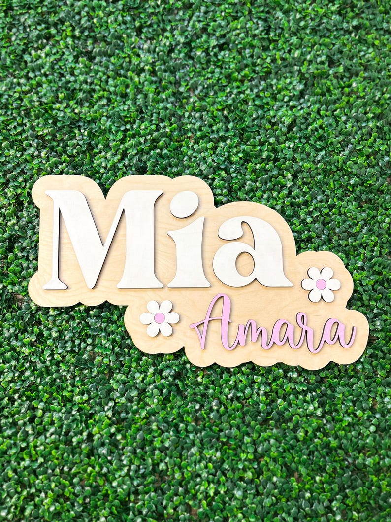 May include: A layered wooden wall sign featuring the name Mia in large white letters and the name Amara in pink cursive script. The design includes two small white flowers with pink centers, all mounted on a light-colored wooden backing board.