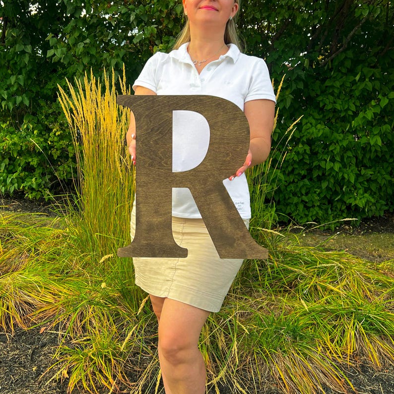 Op de afbeelding: Een grote houten letter R, geverfd in een donkerbruine tint. De letter wordt vastgehouden door een persoon voor een groene, bladerrijke achtergrond.