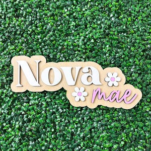 May include: A custom wooden name sign featuring the name Nova Mae in layered lettering. The word Nova is in white, and the word mae is in light pink. Two white flowers with pink centers are attached to the light wood backing board.