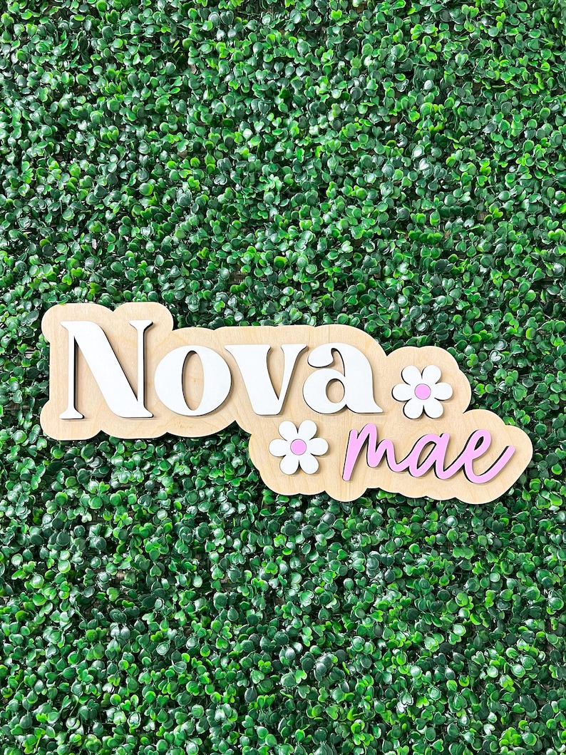 May include: A custom wooden name sign featuring the name Nova Mae in layered lettering. The word Nova is in white, and the word mae is in light pink. Two white flowers with pink centers are attached to the light wood backing board.