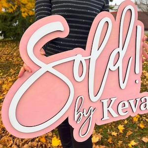 Op de afbeelding: Een groot, op maat gevormd houten bord met een lichtroze achtergrond en witte, verhoogde 3D-letters. Het bord bevat het woord "Sold!" in een groot cursief lettertype, gevolgd door de tekst "by Keva" in een strak, schreefloos lettertype.