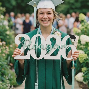 May include: A graduate in a green gown and cap holds a wooden sign that reads "2020 Reagan." The sign has a mint green font. The graduate is smiling, and the background includes greenery and other people.