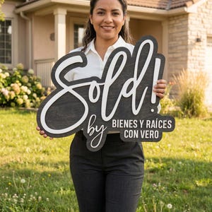 May include: A woman holding a wooden sign that says "Sold!" in large, stylized white lettering against a dark background. The sign also includes the text "by BIENES Y RAÍCES CON VERO" in smaller white letters. The woman is smiling in front of a house.