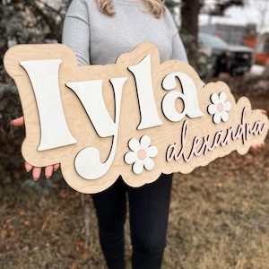 May include: A custom wooden name sign featuring the name Iyla in large white 3D letters and the name alexandra in smaller pink cursive script. The sign includes two white flower cutouts with pink centers, all mounted on a light wood backing.