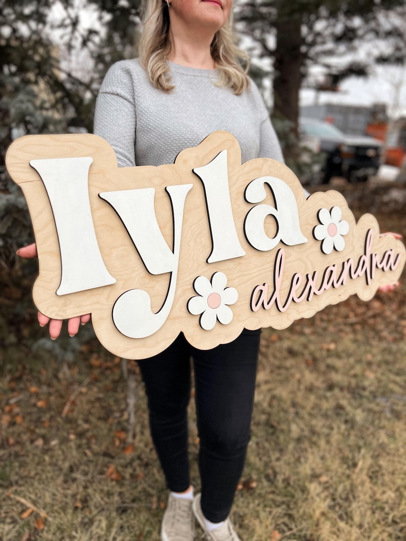May include: A custom wooden name sign featuring the name Iyla in large white 3D letters and the name alexandra in smaller pink cursive script. The sign includes two white flower cutouts with pink centers, all mounted on a light wood backing.