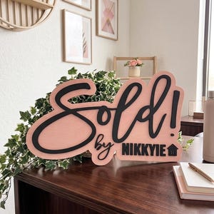 May include: A custom wooden sign featuring a light pink background with raised black lettering that reads Sold! by NIKKYIE with a small house icon. The sign has a smooth finish and is designed for display on a flat surface to announce property sales.