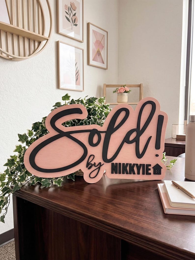 May include: A custom wooden sign featuring a light pink background with raised black lettering that reads Sold! by NIKKYIE with a small house icon. The sign has a smooth finish and is designed for display on a flat surface to announce property sales.