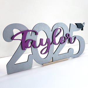 May include: A wooden graduation decoration featuring the year 2025 in gray, with the name "Taylor" in purple script. A small graduation cap is attached to the number 5. The decoration is supported by a wooden base.
