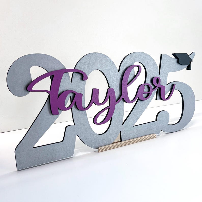 May include: A wooden graduation decoration featuring the year 2025 in gray, with the name "Taylor" in purple script. A small graduation cap is attached to the number 5. The decoration is supported by a wooden base.