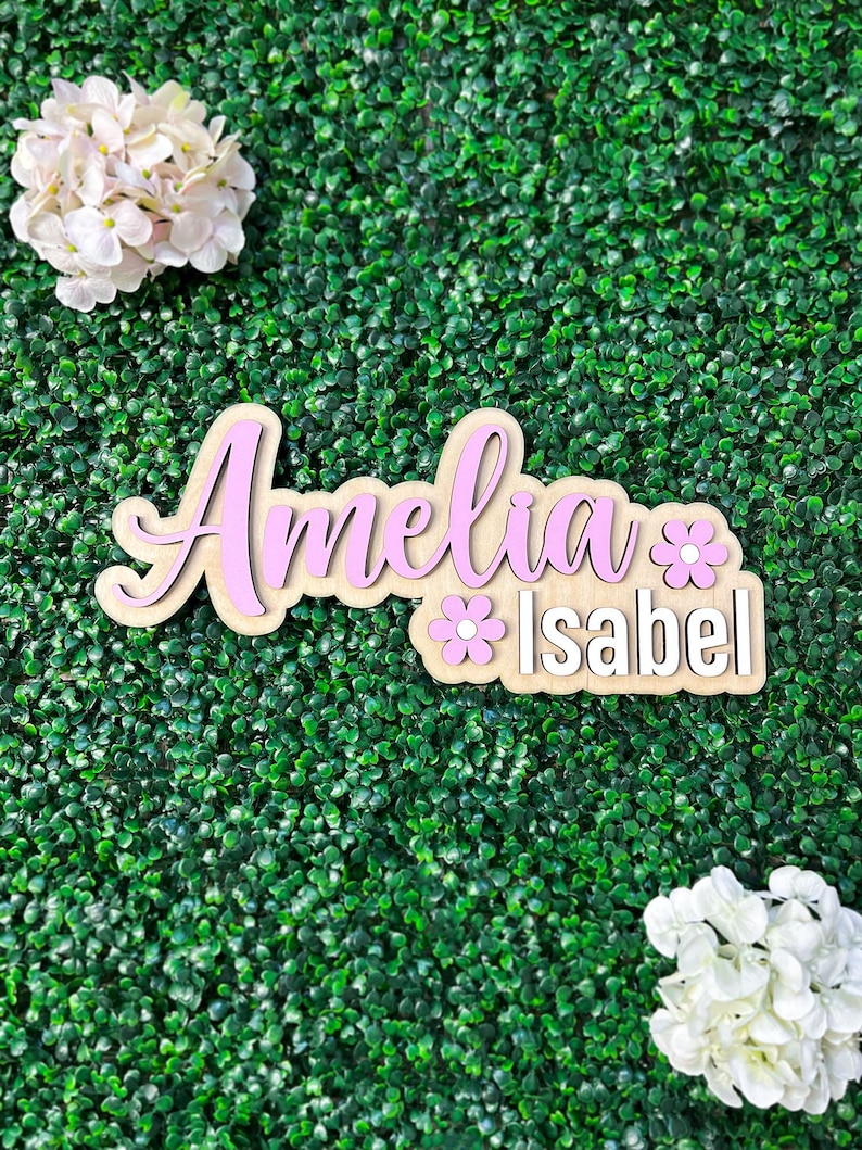 May include: A wooden name sign featuring the name Amelia in large pink script lettering and the name Isabel in white block lettering, accented with two small pink flower cutouts, positioned between two clusters of white hydrangea flowers.