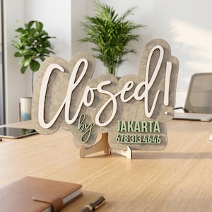 Op de afbeelding: Een decoratief houten bord met een grijze, gemarmerde afwerking, voorzien van de witte scripttekst "Closed!" en de groene tekst "by JAKARTA 678 313 4646". Het bord wordt ondersteund door een kleine, lichtgekleurde houten standaard.