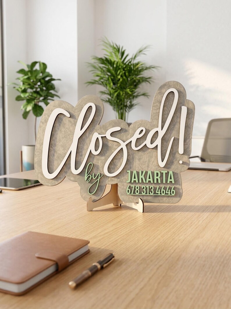 Op de afbeelding: Een decoratief houten bord met een grijze, gemarmerde afwerking, voorzien van de witte scripttekst "Closed!" en de groene tekst "by JAKARTA 678 313 4646". Het bord wordt ondersteund door een kleine, lichtgekleurde houten standaard.