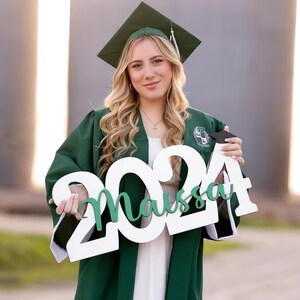 May include: A person wearing a green graduation gown and cap, holding a white sign with the year "2024" and the name "Maissa" in green. The person is smiling, with long blonde hair and a necklace.