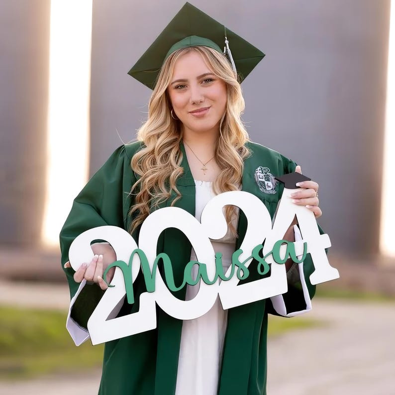 May include: A person wearing a green graduation gown and cap, holding a white sign with the year "2024" and the name "Maissa" in green. The person is smiling, with long blonde hair and a necklace.