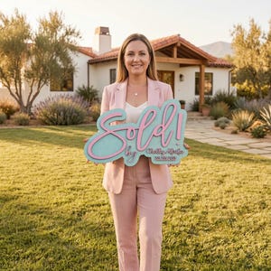 May include: A light pink blazer and matching trousers worn with a white top, holding a teal and pink sign that reads Sold! by Shelby Afraka with a phone number. The sign features a script font for the word Sold and block text for the details.