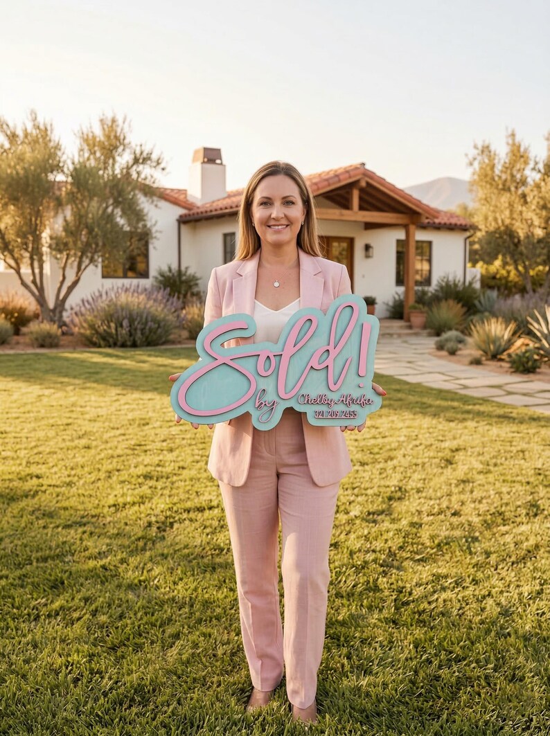 May include: A light pink blazer and matching trousers worn with a white top, holding a teal and pink sign that reads Sold! by Shelby Afraka with a phone number. The sign features a script font for the word Sold and block text for the details.