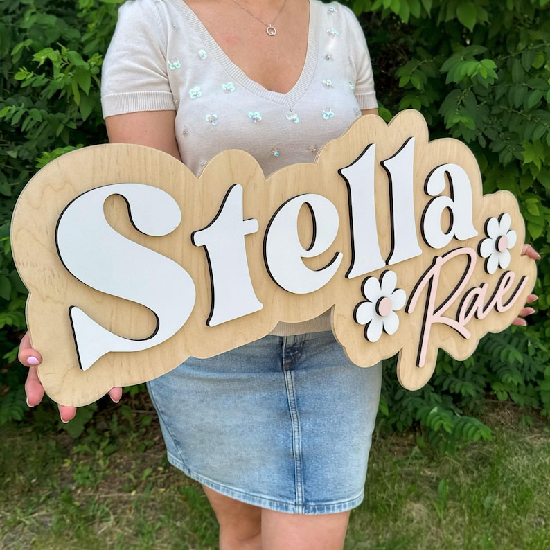 May include: A custom wooden name sign featuring the name Stella Rae in layered white and light pink lettering. The sign has a natural wood backing with a scalloped edge and includes two white flower accents with light pink centers on the right side.