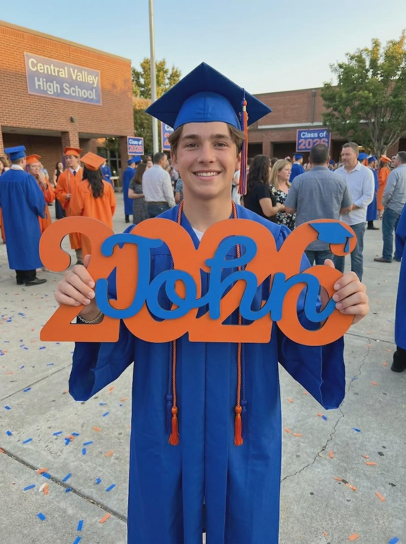 May include: A smiling graduate in a blue cap and gown holds a sign that reads "2025 John" in orange and blue. The background shows a high school building and other graduates in orange and blue gowns.