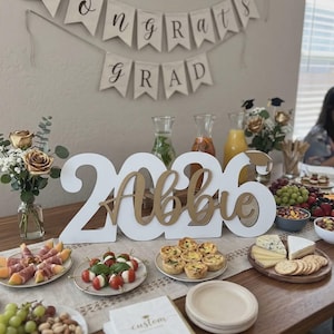 May include: A graduation party table setting with a banner that reads "Congrats Grad." The table features a large "2026 Abbie" sign, various appetizers, drinks, and floral arrangements. Napkins with the word "custom" are also visible.