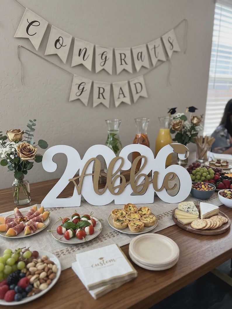 May include: A graduation party table setting with a banner that reads "Congrats Grad." The table features a large "2026 Abbie" sign, various appetizers, drinks, and floral arrangements. Napkins with the word "custom" are also visible.