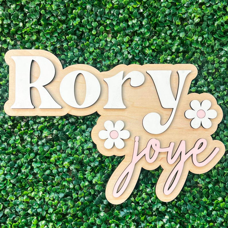 May include: A wooden wall sign featuring the name Rory in large white serif letters and the name joye in smaller light pink cursive script. Two white flowers with light pink centers are attached to the light-colored wooden backing board.