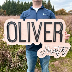 May include: A custom wooden name sign featuring the name OLIVER in large, bold, black sans-serif capital letters mounted on a light-colored wood backing, with the name Hunter in white cursive script lettering positioned below it.