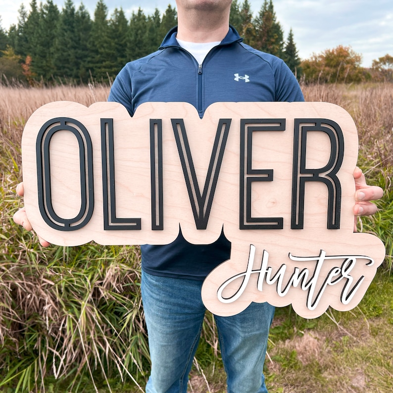 May include: A custom wooden name sign featuring the name OLIVER in large, bold, black sans-serif capital letters mounted on a light-colored wood backing, with the name Hunter in white cursive script lettering positioned below it.