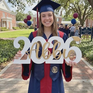 May include: A graduate in a navy blue cap and gown with red accents holds a white sign with "2026 Abbie" in gold lettering. The sign includes a graduation cap icon. The background shows a school building and balloons.