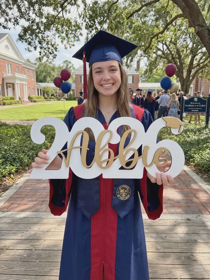 May include: A graduate in a navy blue cap and gown with red accents holds a white sign with "2026 Abbie" in gold lettering. The sign includes a graduation cap icon. The background shows a school building and balloons.
