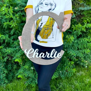 May include: A wooden letter C with the name "Charlie" written in white paint. The letter is being held by a person wearing a white shirt with a yellow vest design and black trousers.