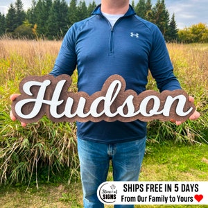 May include: A wooden sign with the name "Hudson" in a layered design. The top layer features white cursive lettering, while the bottom layer is a dark brown wood. The sign is held in front of a natural outdoor background.