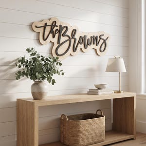 May include: A wall-mounted wooden sign featuring the text "the Browns" in black script lettering layered over a light-coloured, cloud-shaped wooden backing. The sign is displayed above a wooden console table with a plant, lamp, and basket.