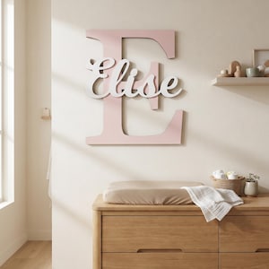 May include: A large, light pink wooden letter E mounted on a wall, featuring the name Elise in white cursive script layered over the centre. The letter E is block-style, while the name Elise is in a flowing, connected font, creating a dimensional effect.