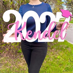 May include: A wooden graduation sign with the year "2023" and the name "Kendel" in pink script. The sign also features a pink graduation cap. The sign is held in front of a person, with a background of green grass and foliage.