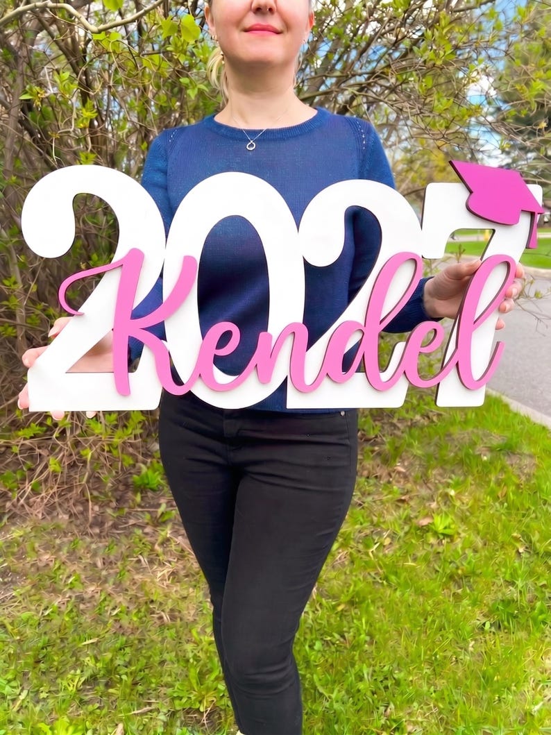 May include: A wooden graduation sign with the year "2023" and the name "Kendel" in pink script. The sign also features a pink graduation cap. The sign is held in front of a person, with a background of green grass and foliage.