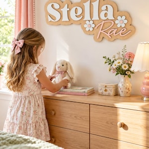 May include: A wooden wall sign featuring the name Stella Rae in white and pink layered lettering, accented with two white flowers with pink centers. The sign is mounted on a light wood-toned backing with a cloud-like, scalloped edge shape.