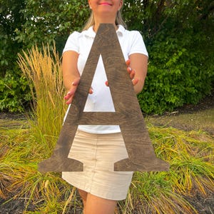 Op de afbeelding: Een grote, houten letter A, gebeitst in een donkerbruine kleur. De letter wordt vastgehouden door een persoon voor een grasachtige achtergrond.