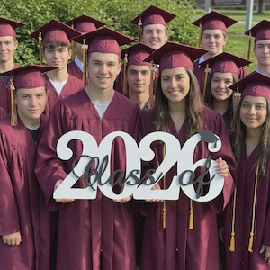 May include: A group of graduates wearing maroon gowns and caps, holding a sign that reads "Class of 2026". The sign is white with black lettering. The graduates are smiling and posing for a photo.
