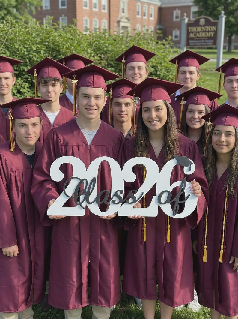May include: A group of graduates wearing maroon gowns and caps, holding a sign that reads "Class of 2026". The sign is white with black lettering. The graduates are smiling and posing for a photo.