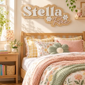 May include: A custom wooden wall sign featuring the name Stella Rae in white and pink layered lettering, accented with two white flower cutouts. The sign is mounted on a light wood backing with a scalloped edge, displayed above a wooden bed frame.