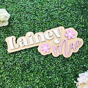 May include: A custom wooden sign featuring the name Lainey Mae in layered lettering. The word Lainey is in white, and the word Mae is in pink. Two light pink flowers with white centers are attached to the light-colored wooden base board.