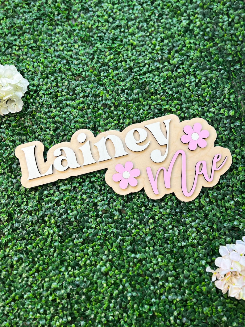 May include: A custom wooden sign featuring the name Lainey Mae in layered lettering. The word Lainey is in white, and the word Mae is in pink. Two light pink flowers with white centers are attached to the light-colored wooden base board.