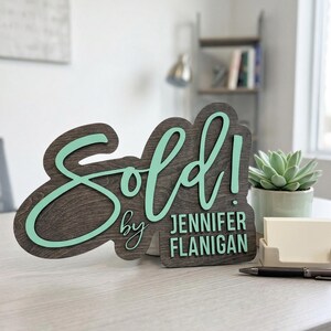 Op de afbeelding: Een donkerhouten bord met een licht mintgroene 3D-uitgesneden tekst die Sold! leest en de tekst by JENNIFER FLANIGAN in een bijpassend mintgroen lettertype. Het bord heeft een gelaagd ontwerp met een donker gebeitste houten achterkant.