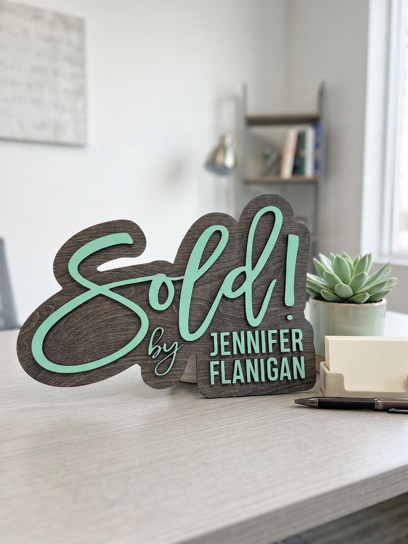 Op de afbeelding: Een donkerhouten bord met een licht mintgroene 3D-uitgesneden tekst die Sold! leest en de tekst by JENNIFER FLANIGAN in een bijpassend mintgroen lettertype. Het bord heeft een gelaagd ontwerp met een donker gebeitste houten achterkant.