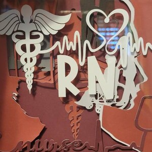 May include: A framed papercraft artwork with a red and white color scheme. The artwork features a stylized medical symbol, a heartbeat, and the letters "RN" in white. The word "nurse" is also visible in the bottom left corner.