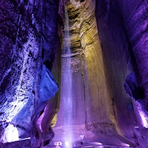 May include: A large waterfall cascades down a cavernous rock formation, illuminated in shades of purple and yellow. The water appears to be flowing from a high opening in the ceiling of the cave.
